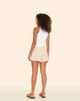 A person with curly hair is against a beige background, wearing a crisp white sleeveless top, Xirena's Carlyn Short in peach with an elasticized waist, and brown sandals. They stand slightly turned away, showing their side and back.