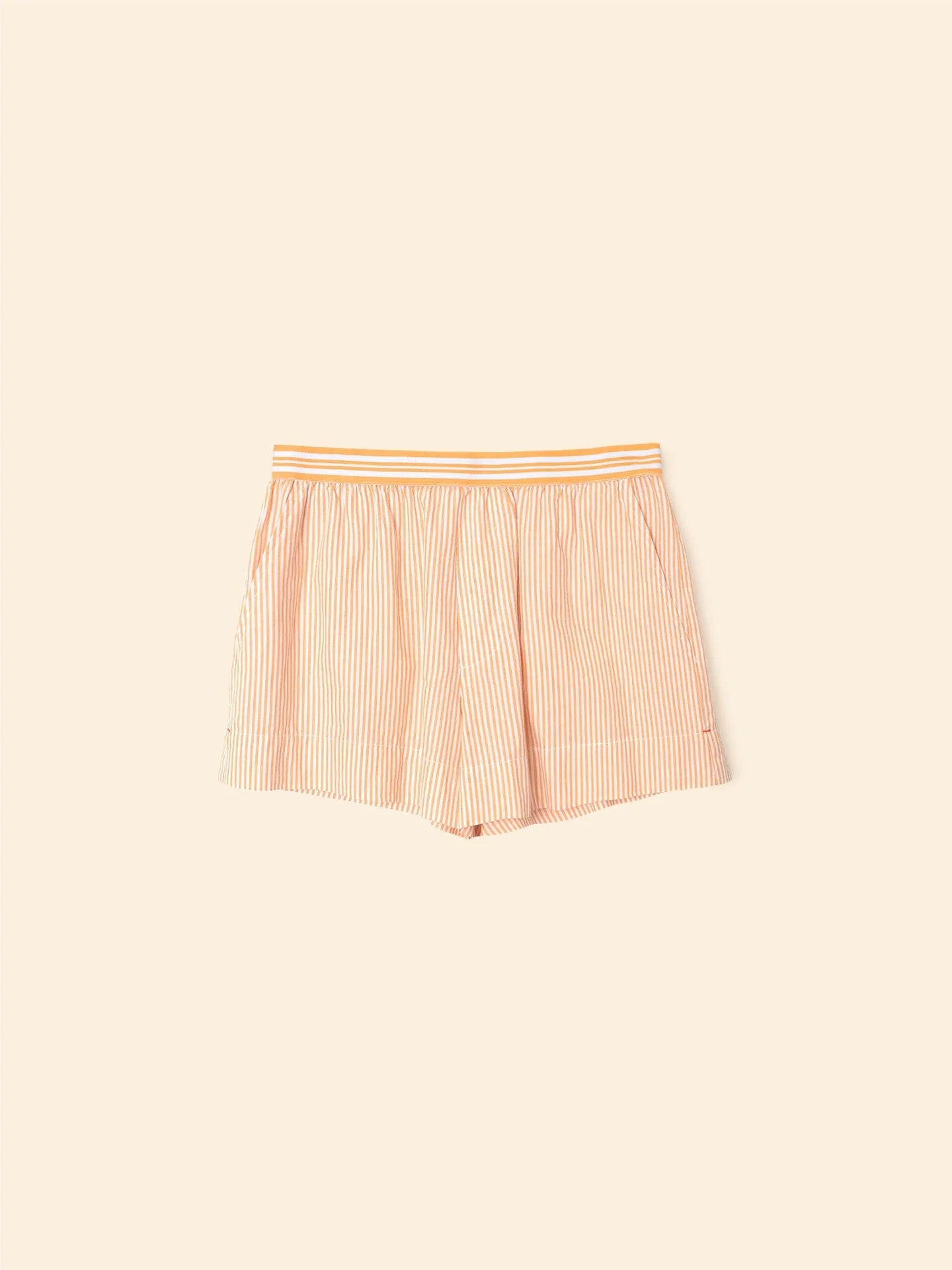 The Carlyn Short by Xirena features orange and white pencil stripes with a thicker orange stripe, an elasticized waistband, and is made from crisp cotton. The shorts are shown against a light beige background.