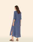 A woman with brown hair wears the Xirena Boden Dress, a tailored long blue cotton poplin shirtdress with rolled-up sleeves and side slits, paired with brown sandals. She's standing against a neutral beige background.