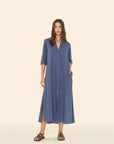 A woman wears the Xirena Boden Dress—a loose, long-sleeve blue cotton poplin shirtdress with side pockets and front slits—styled with brown sandals, standing against a plain beige background.