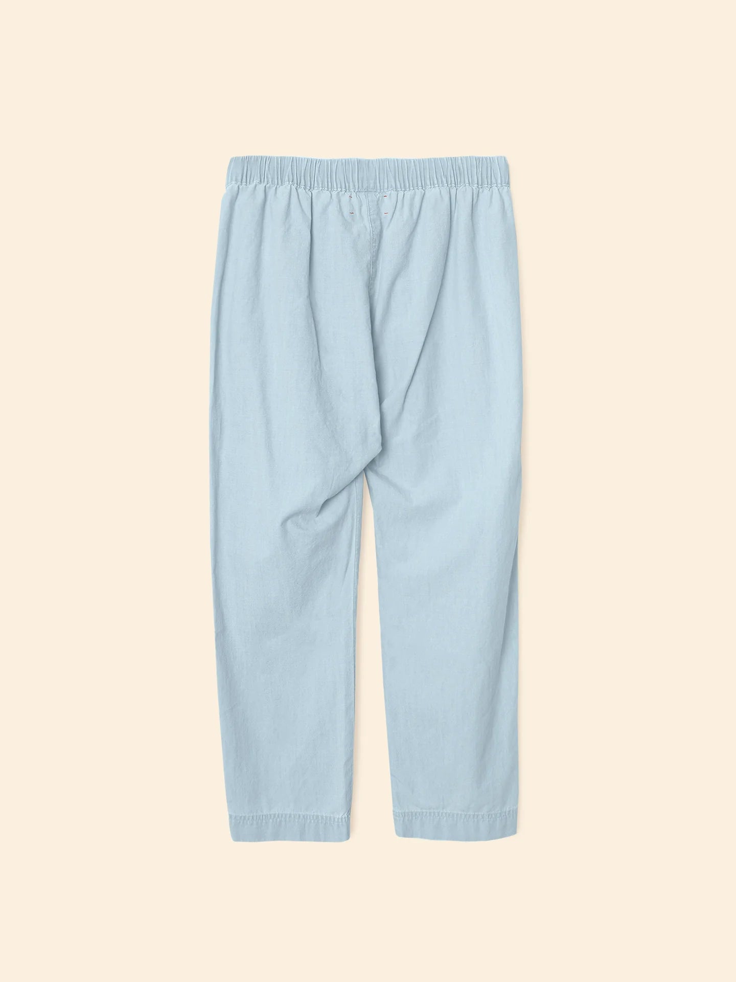 The Xirena Draper Pant is an ankle-length, light blue chambray style with a relaxed fit, elastic waistband, and side seam pockets. Shown flat on a beige background.