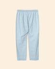 The Xirena Draper Pant is an ankle-length, light blue chambray style with a relaxed fit, elastic waistband, and side seam pockets. Shown flat on a beige background.