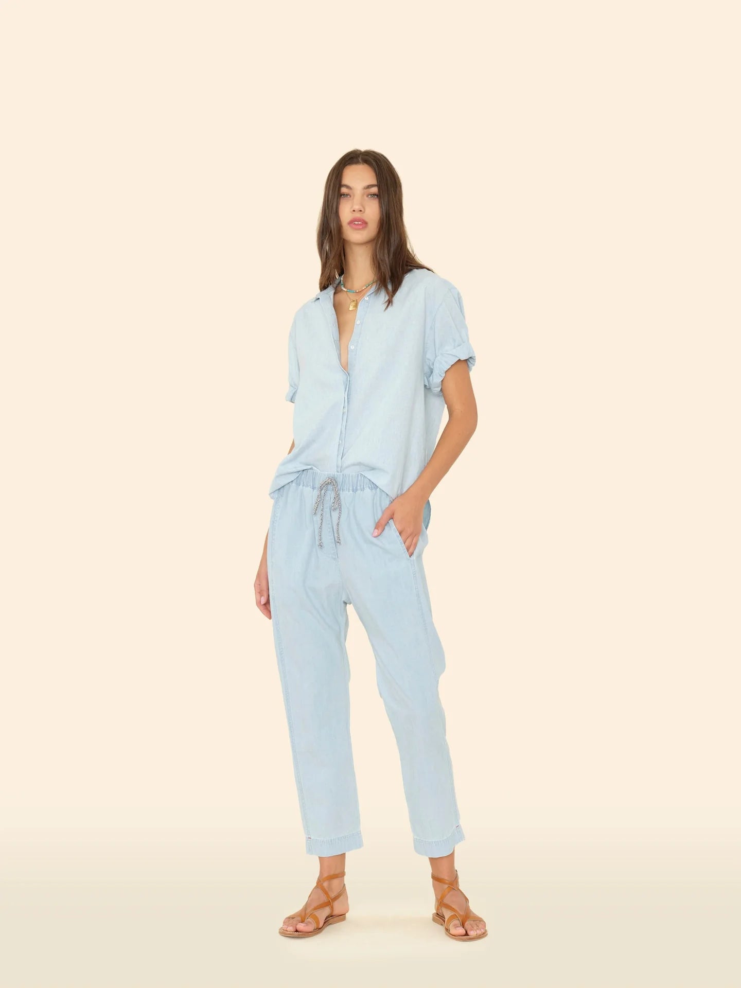 A woman stands against a beige background wearing Xirena’s Draper Pant, a light blue lightweight cotton chambray drawstring waist pant with side seam pockets, paired with a matching short-sleeve button-up shirt and tan sandals.