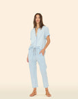 A woman stands against a beige background wearing Xirena’s Draper Pant, a light blue lightweight cotton chambray drawstring waist pant with side seam pockets, paired with a matching short-sleeve button-up shirt and tan sandals.