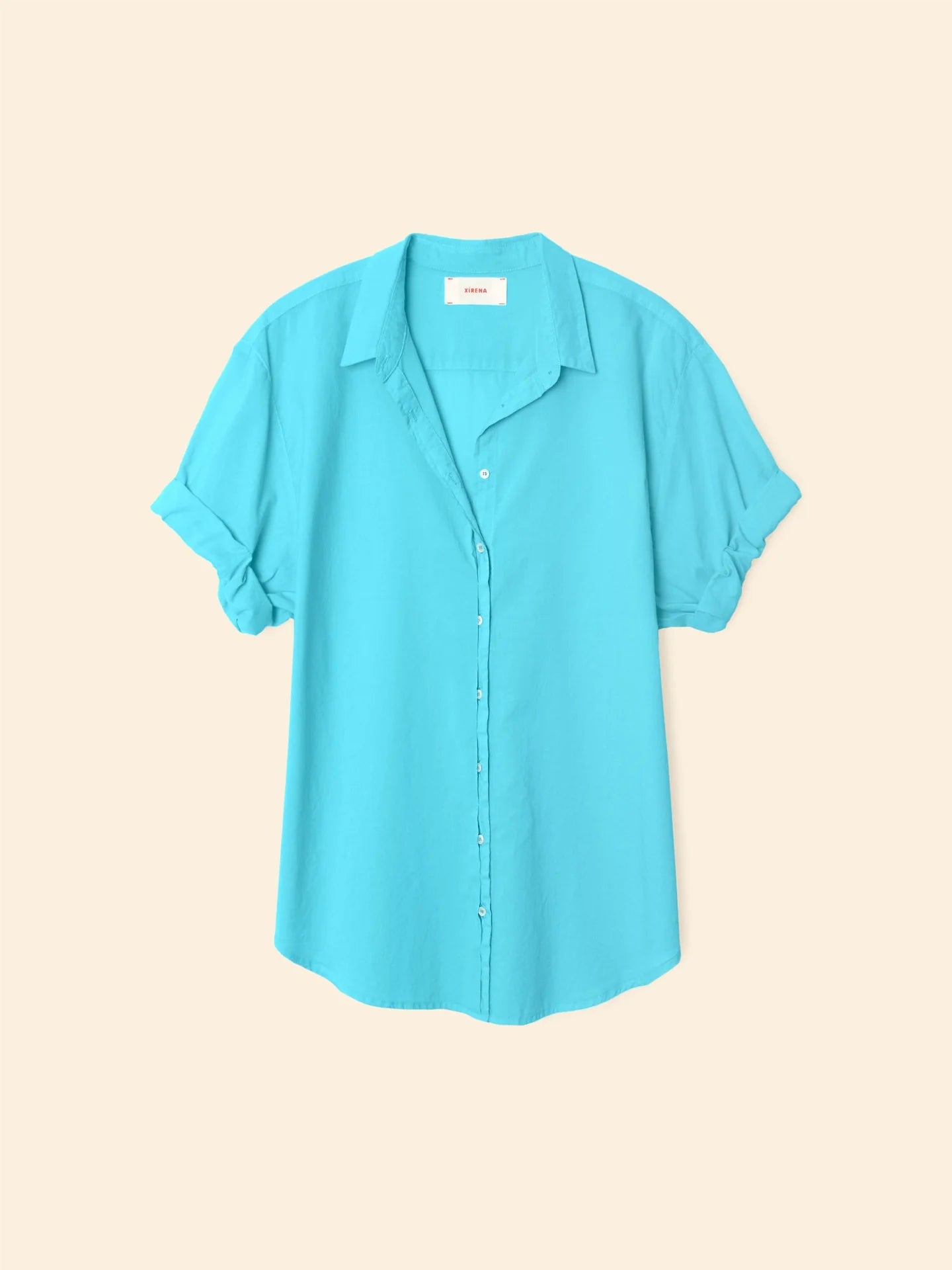 The Xirena Channing Shirt S25 features a light blue cotton chambray, short rolled sleeves, and a classic collar. Shown against a beige background, the shirt offers a relaxed, lightweight feel.