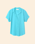 The Xirena Channing Shirt S25 in turquoise, crafted from lightweight cotton chambray with rolled cuffs and a relaxed "boy meets girl" vibe, is displayed against a plain beige background.