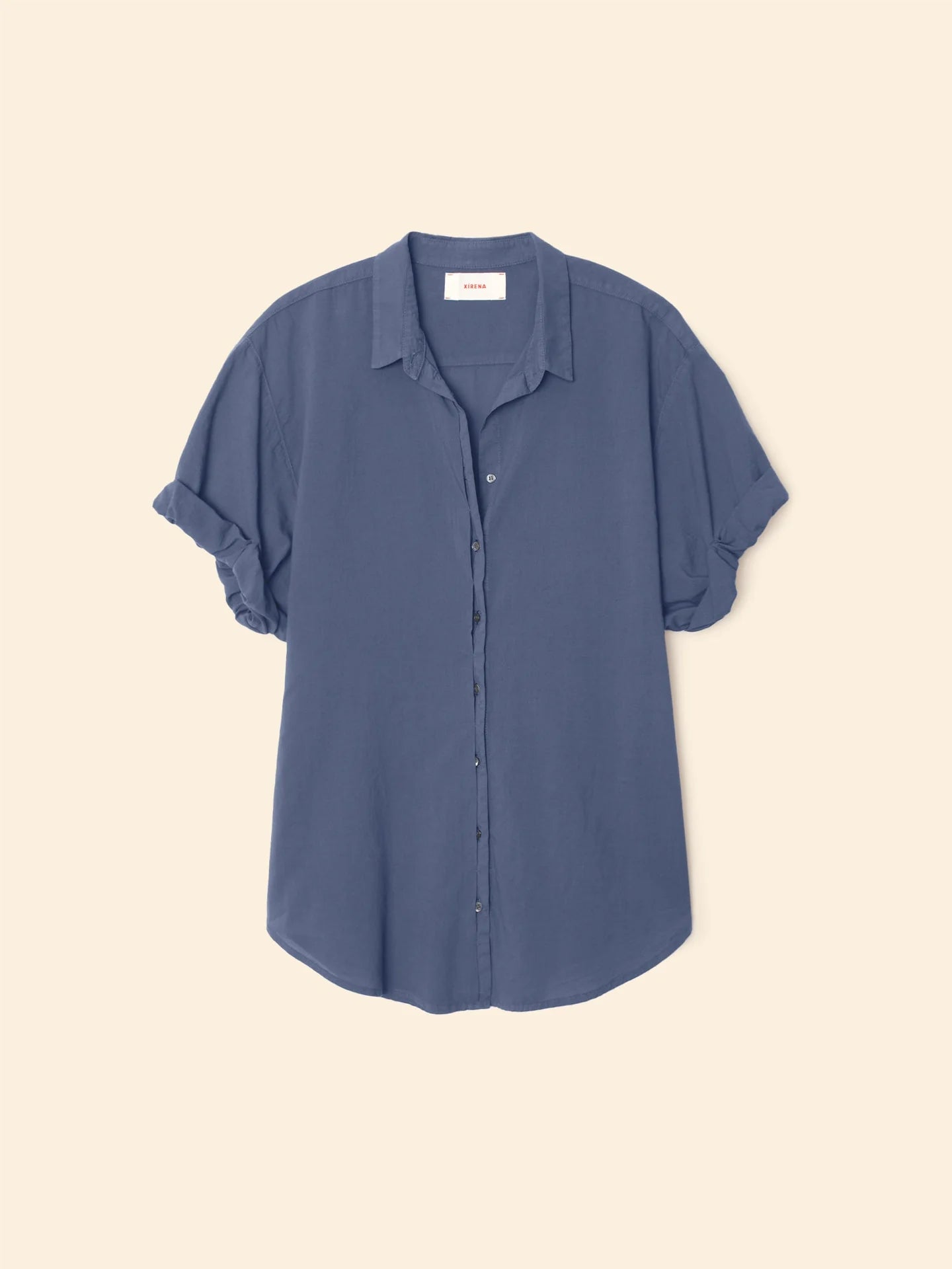 The Xirena Channing Shirt S25 is a short-sleeve, light blue cotton chambray button-up with rolled cuffs, classic collar, and a relaxed fit, shown against a beige background.