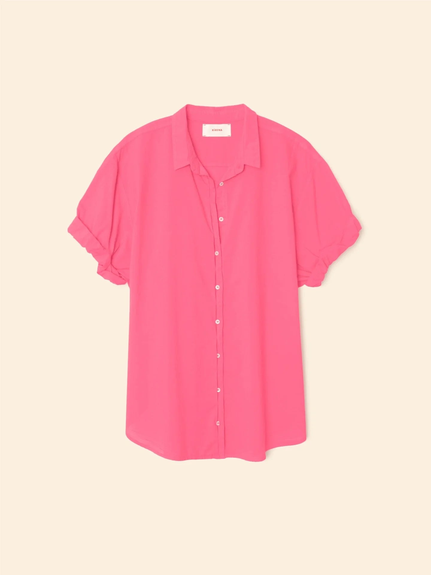 The Xirena Channing Shirt S25, a bright pink cotton chambray shirt with a collar and rolled cuffs, is displayed against a light beige background.