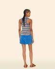 A woman with braided hair stands against a beige background, wearing the Xirena Shayden Short in bright blue and a striped sleeveless top with brown sandals.