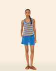A woman with braided hair stands against a light beige background, wearing the Xirena Shayden Short in bright blue with an elasticized waist, a cotton blend striped tank top, and brown sandals. She looks at the camera with one hand in her pocket.