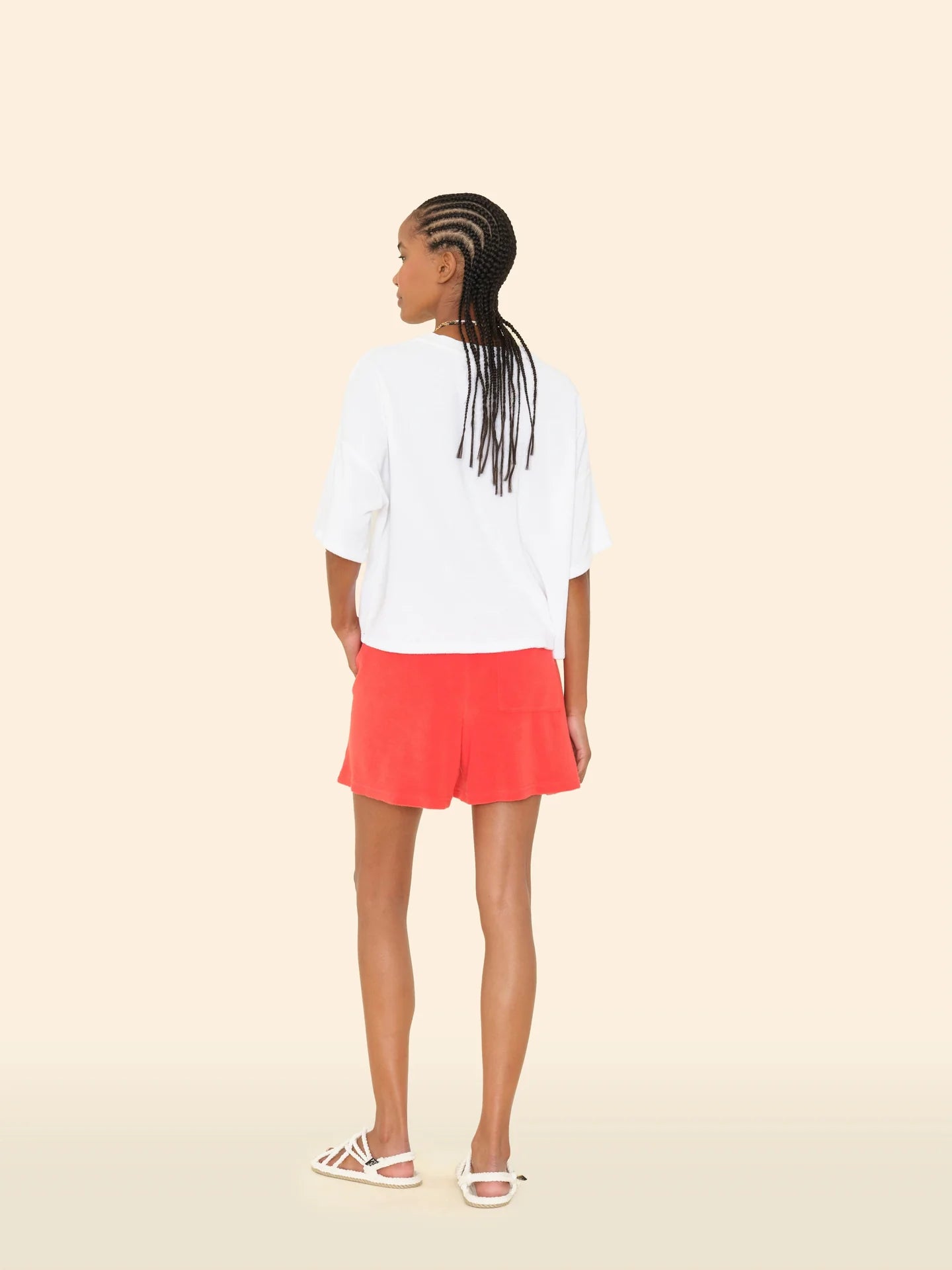 A person with braided hair faces away, wearing a loose white T-shirt, Xirena Shayden Shorts in bright red with an elastic waist, and white sandals, set against a plain beige background.