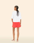 A person with braided hair faces away, wearing a loose white T-shirt, Xirena Shayden Shorts in bright red with an elastic waist, and white sandals, set against a plain beige background.