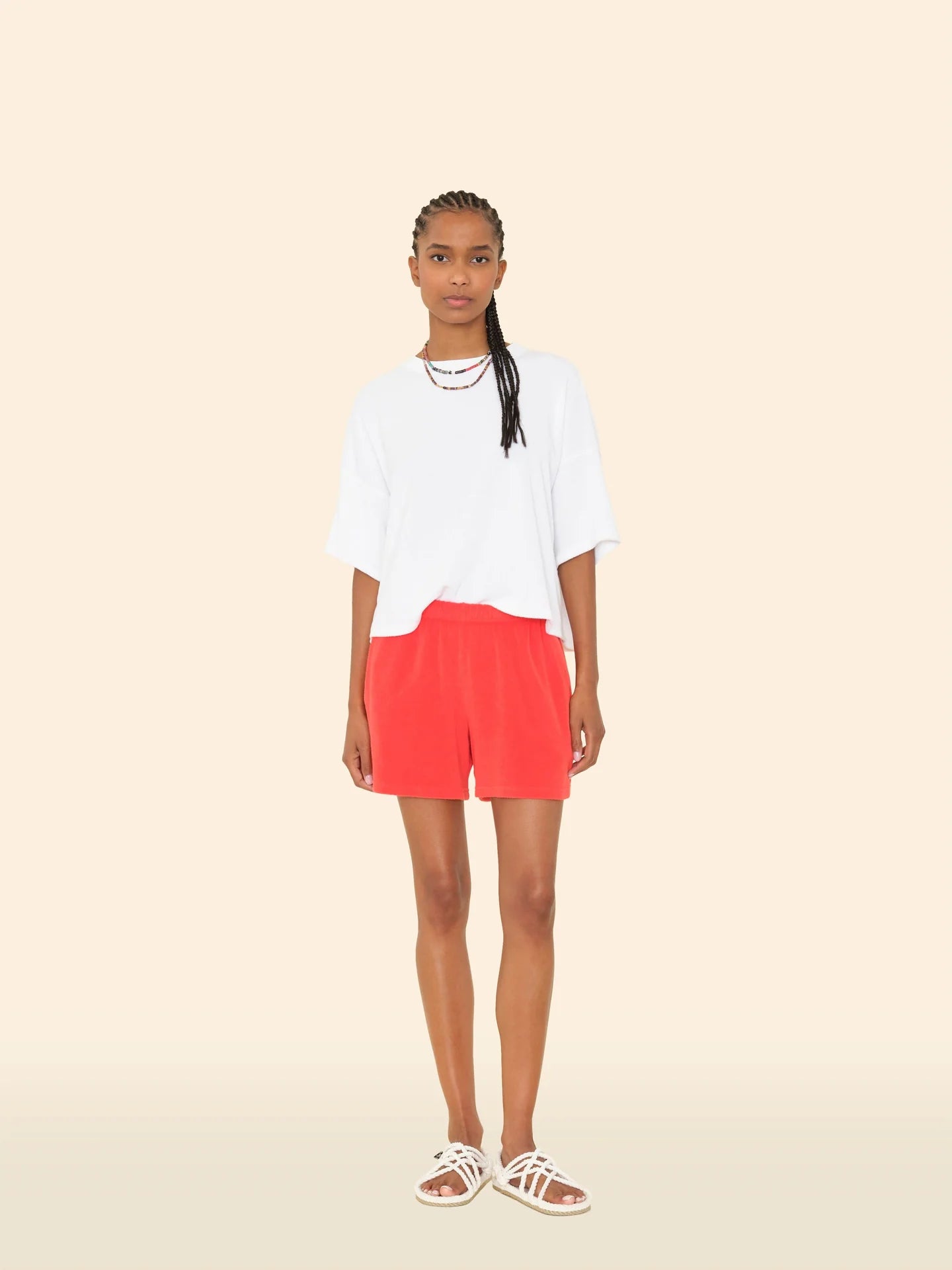 A person with long braided hair and a neutral expression stands against a beige background, wearing the Xirena Shayden Short in bright red, a loose white T-shirt, white strappy sandals, and layered necklaces.