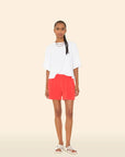 A person with long braided hair and a neutral expression stands against a beige background, wearing the Xirena Shayden Short in bright red, a loose white T-shirt, white strappy sandals, and layered necklaces.