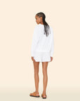 A woman with braided hair stands facing away, wearing a white cotton blend long-sleeve top and Xirena’s Shayden Short, featuring an elasticized waist, paired with tan sandals against a plain beige background.