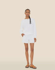 A woman with braided hair poses against a beige background in a white long-sleeve shirt, Xirena Shayden Shorts with an elastic waist, brown sandals, and a brown necklace. One hand is in her pocket.