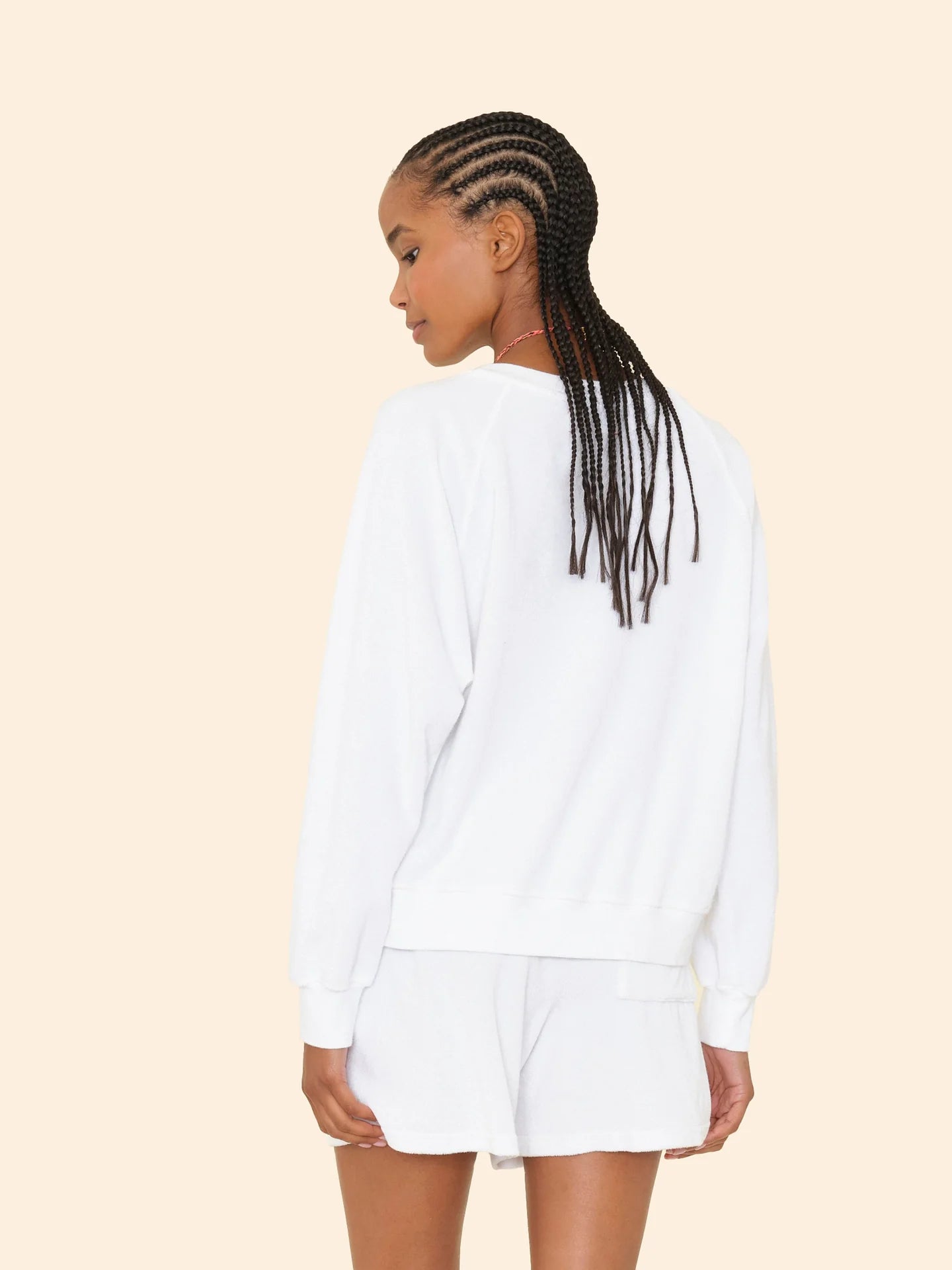 A woman with long, braided hair stands facing away, wearing the Xirena Hopper Sweatshirt and matching white shorts in a cotton-blend terry fabric against a light beige background.