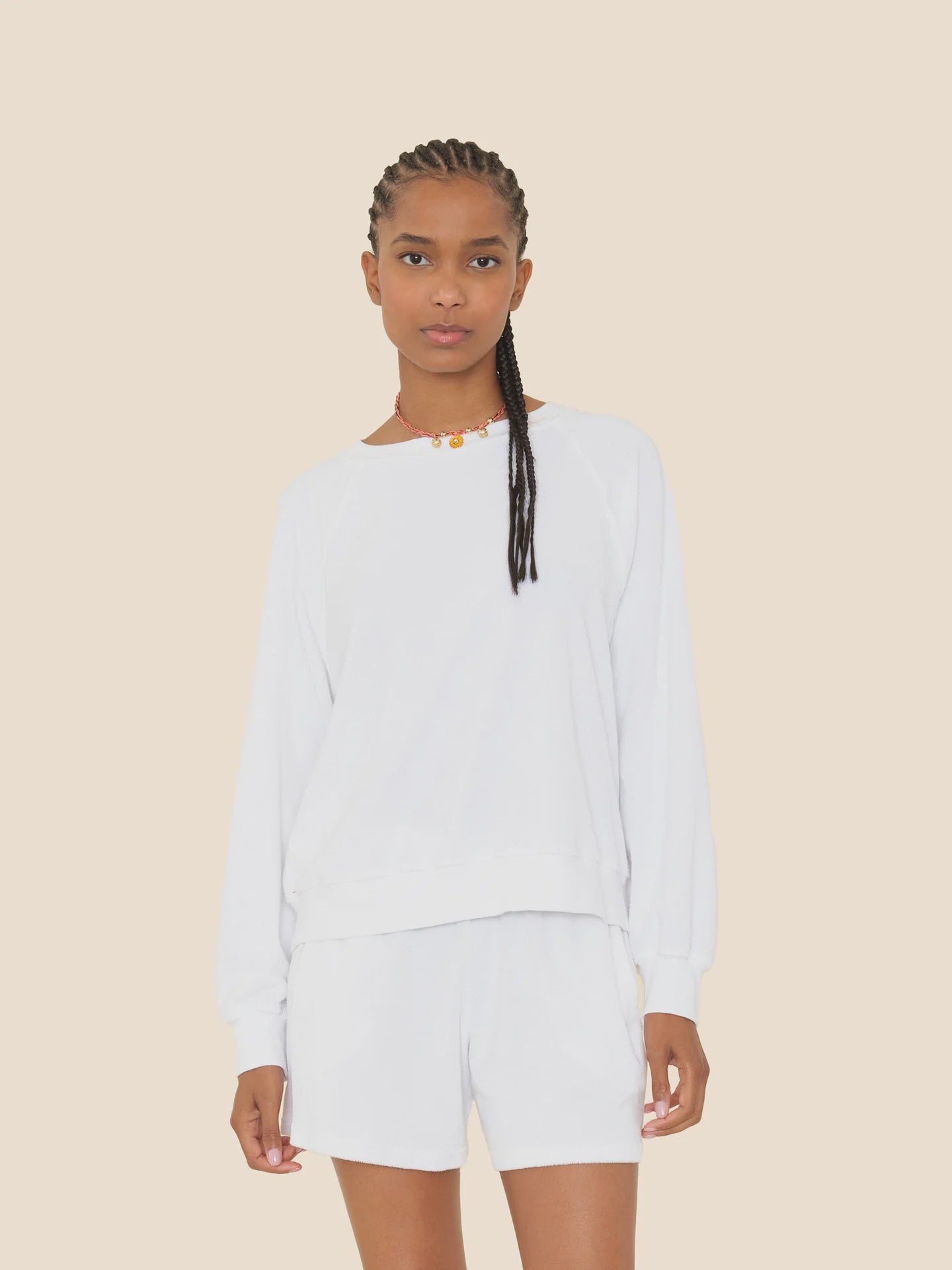 A woman with braided hair models the Xirena Hopper Sweatshirt, a cotton blend piece, paired with matching white shorts against a plain beige background. Made in Los Angeles.