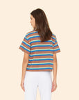 A woman with long brown hair, seen from behind, wears the Xirena Paulina Tee—a striped cotton jersey in blue, white, and yellow with a contrast ringer neck—paired with white pants against a plain beige background.