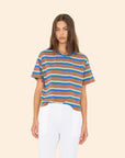 A woman with long brown hair wears the Xirena Paulina Tee in blue and white striped cotton jersey with a contrast ringer neck, paired with white pants, standing against a plain beige background.