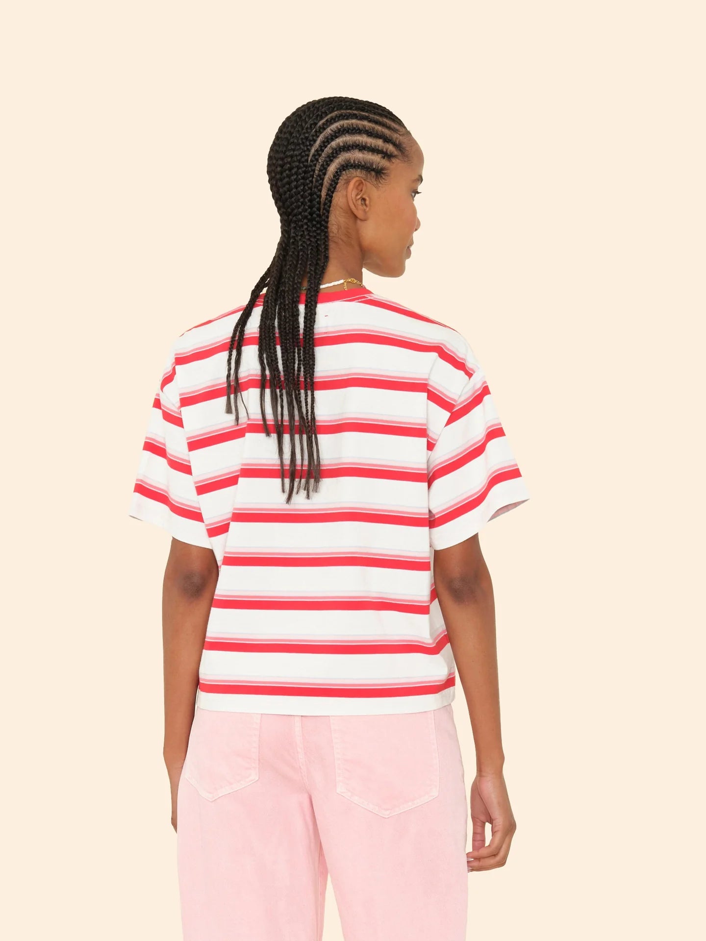 A person with braided hair is seen from behind, wearing a striped cotton Paulina Tee by Xirena with a contrast ringer neck and light pink pants against a beige background.