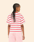A person with braided hair is seen from behind, wearing a striped cotton Paulina Tee by Xirena with a contrast ringer neck and light pink pants against a beige background.