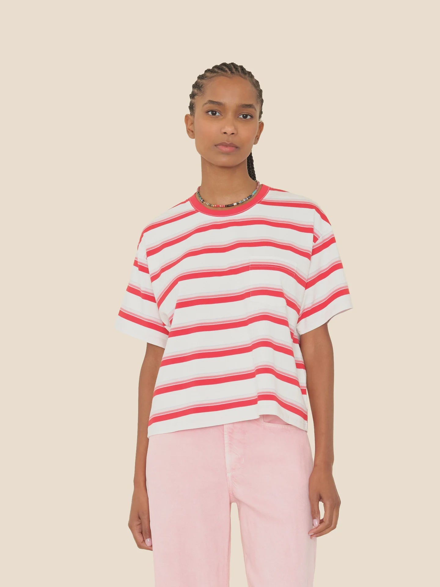 A person with braided hair wears a loose white Paulina Tee by Xirena with red horizontal stripes and light pink pants, standing against a plain beige background.