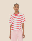 A person with braided hair wears a loose white Paulina Tee by Xirena with red horizontal stripes and light pink pants, standing against a plain beige background.