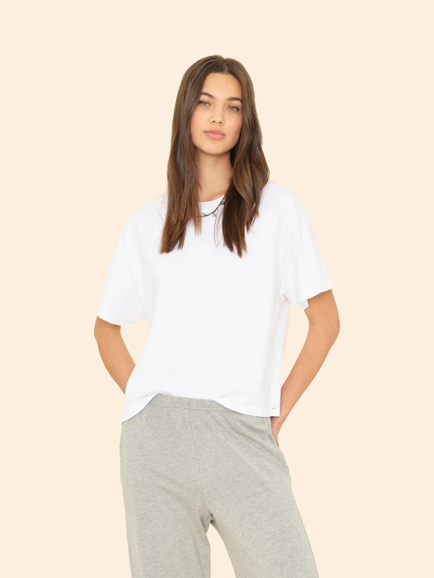 A woman with long brown hair wears the Xirena Palmer Tee, a loose cotton jersey wide-sleeve top, and light gray sweatpants, standing against a plain beige background with a relaxed expression.