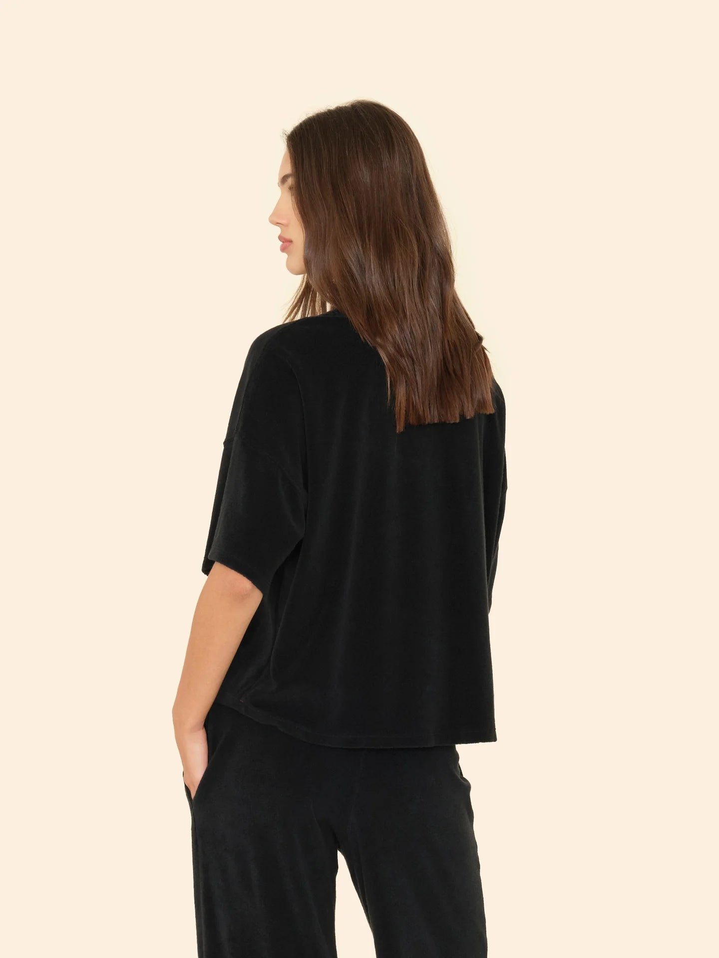 A woman with long brown hair is shown from the side and back, wearing a loose-fitting Xirena Roe Top and black pants, her right hand in her pocket, standing against a plain beige background.