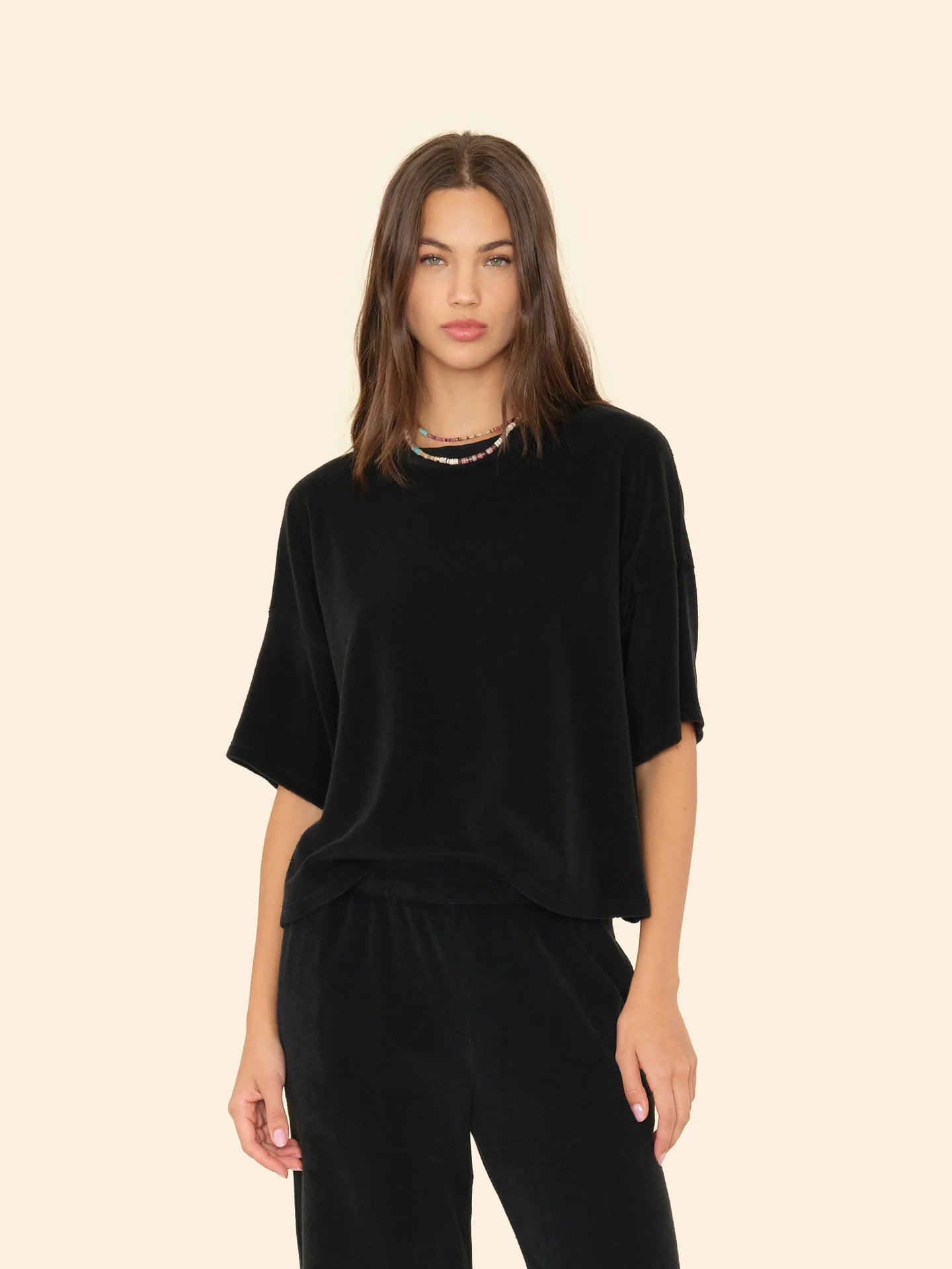 A woman with straight brown hair wears the Xirena Roe Top, a loose-fitting black cotton-blend sweatshirt with wide sleeves, paired with matching pants and a multicolored beaded necklace, standing against a plain beige background.