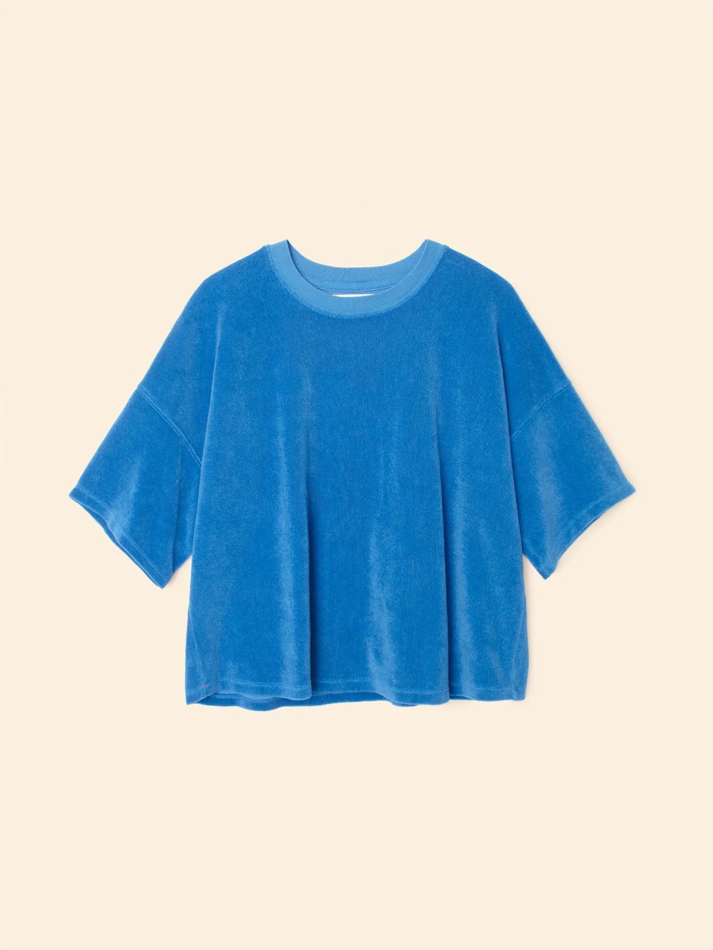 The Roe Top by Xirena is a loose-fitting, short-sleeved blue velour T-shirt featuring a darker blue crew neckline, made from a soft cotton blend, shown on a plain beige background.