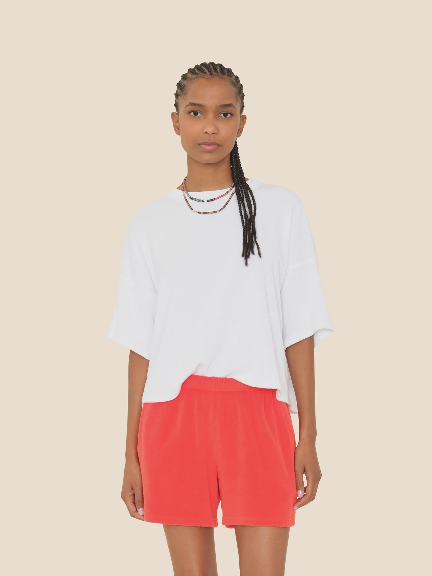 A person with braided hair wears the Roe Top by Xirena, a loose white cotton blend T-shirt, and bright red shorts, standing against a plain beige background.