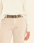 A person wears the Xirena Francoise Belt—a brown leather belt with large gold grommets and an antique brass buckle—over beige pants and a cream sweater. Only the lower torso and one hand are visible.