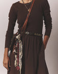 A person wearing a long-sleeve brown dress with the Xirena Francoise Belt—an adjustable black leather belt with an antique brass buckle—layered necklaces, a brown crossbody bag, and a patterned tassel scarf attached to the bag.