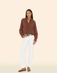 A woman poses against a beige background wearing the Xirena Amelie Shirt—a brown long sleeve button-up with subtle ruffle details—paired with white pants, a patterned belt, and leopard print flats, one hand in her pocket.