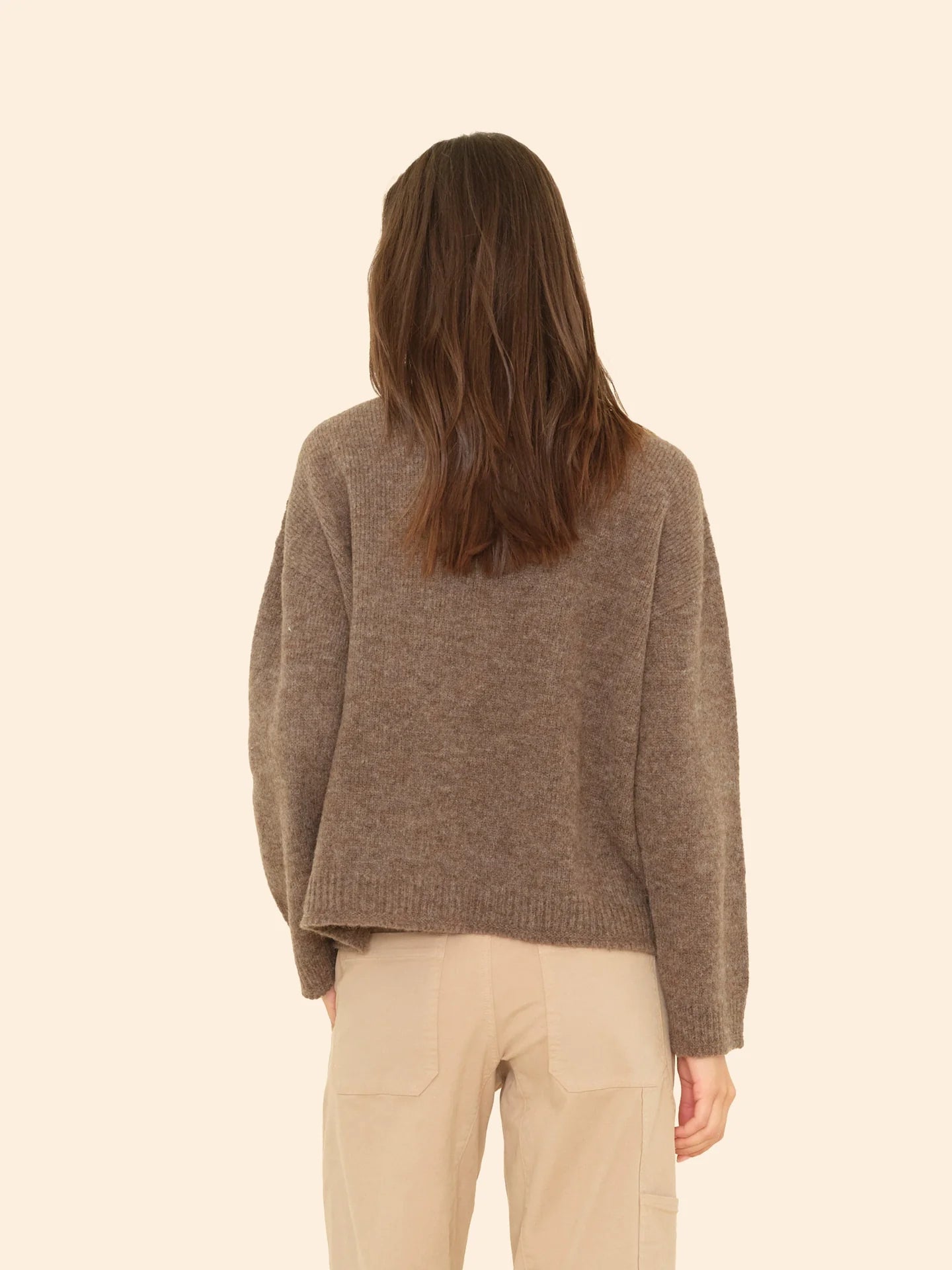 A woman with long brown hair, seen from behind, wears the Xirena Cameron Sweater and beige pants, standing against a plain light beige background.