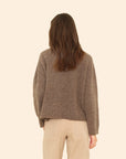 A woman with long brown hair, seen from behind, wears the Xirena Cameron Sweater and beige pants, standing against a plain light beige background.