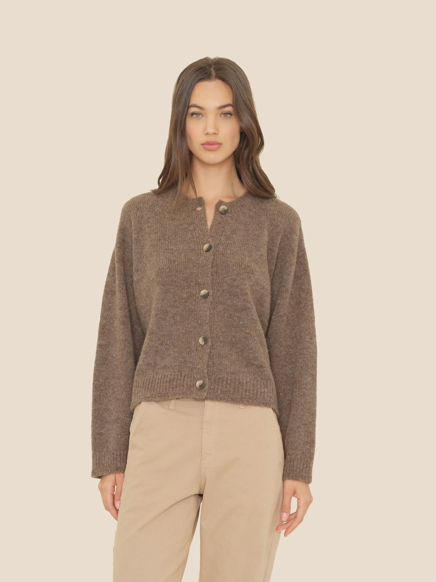 A woman with long brown hair wears the Xirena Cameron Sweater, featuring faux horn buttons, paired with beige pants against a plain light beige background.