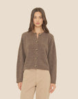 A woman with long brown hair wears the Xirena Cameron Sweater, featuring faux horn buttons, paired with beige pants against a plain light beige background.