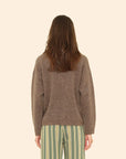 A person with long brown hair, seen from behind, wears the Xirena Ethan Sweater and green pants with vertical white and yellow stripes, standing against a beige background.