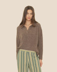 A woman wears the brown Ethan Sweater by Xirena, featuring buttons, paired with green pants with vertical cream and yellow stripes, standing against a plain beige background.