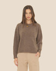 A woman with long brown hair models the Xirena Julien Sweater—a loose merino wool pullover—paired with beige pants, standing against a plain light background with one hand in her pocket.