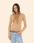 A woman with straight, dark hair stands against a beige background, wearing a tan sweater and light blue jeans, accessorized with the ultra-soft Myla Scarf by Xirena draped over her shoulders and a black belt with silver eyelets.