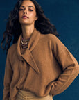 A woman with long wavy dark hair wears a brown loose-collared sweater, beige skirt, layered beaded necklace, and the ultra-soft Myla Scarf by Xirena, standing against a dark blue background.