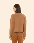 A woman with shoulder-length brown hair faces away, wearing the ultra-soft Xirena Lela Sweater and matching suede pants against a light beige background.