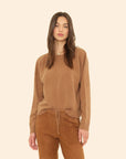 A woman with long brown hair models the Xirena Lela Sweater and matching tan pants, standing against a plain beige background.