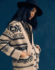 A woman wears the Xirena Vail Cardigan, layered necklaces, and a black wide-brim hat while standing against a dark blue background with her hands in her pockets, gazing downward in thought.
