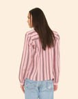 A woman with long straight brown hair stands with her back to the camera against a beige background, wearing the Xirena Sabina Top—a drapey pink striped blouse—paired with light blue jeans.