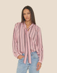 A woman wears the Sabina Top by Xirena—a drapey viscose blouse with black vertical stripes, a front tie, and deep V-neck—paired with light blue jeans, standing against a plain beige background.